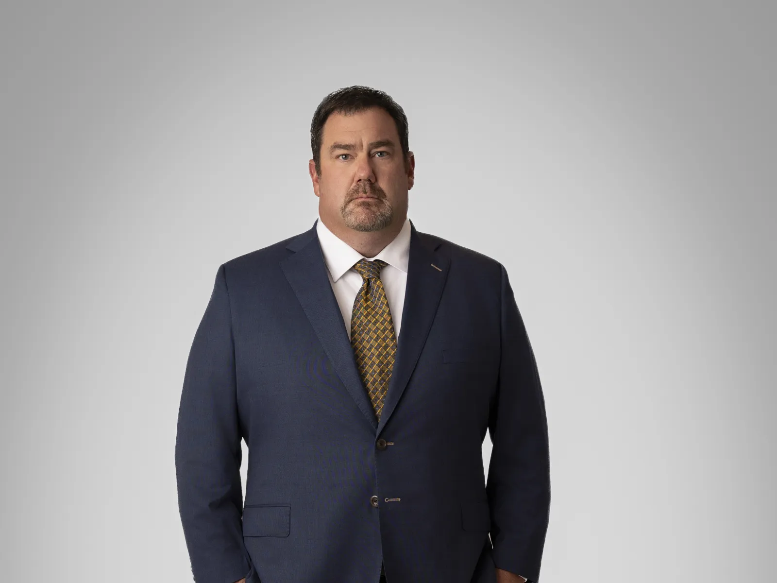 Confident man in a navy suit and tie, standing with hands in pockets against a neutral background.