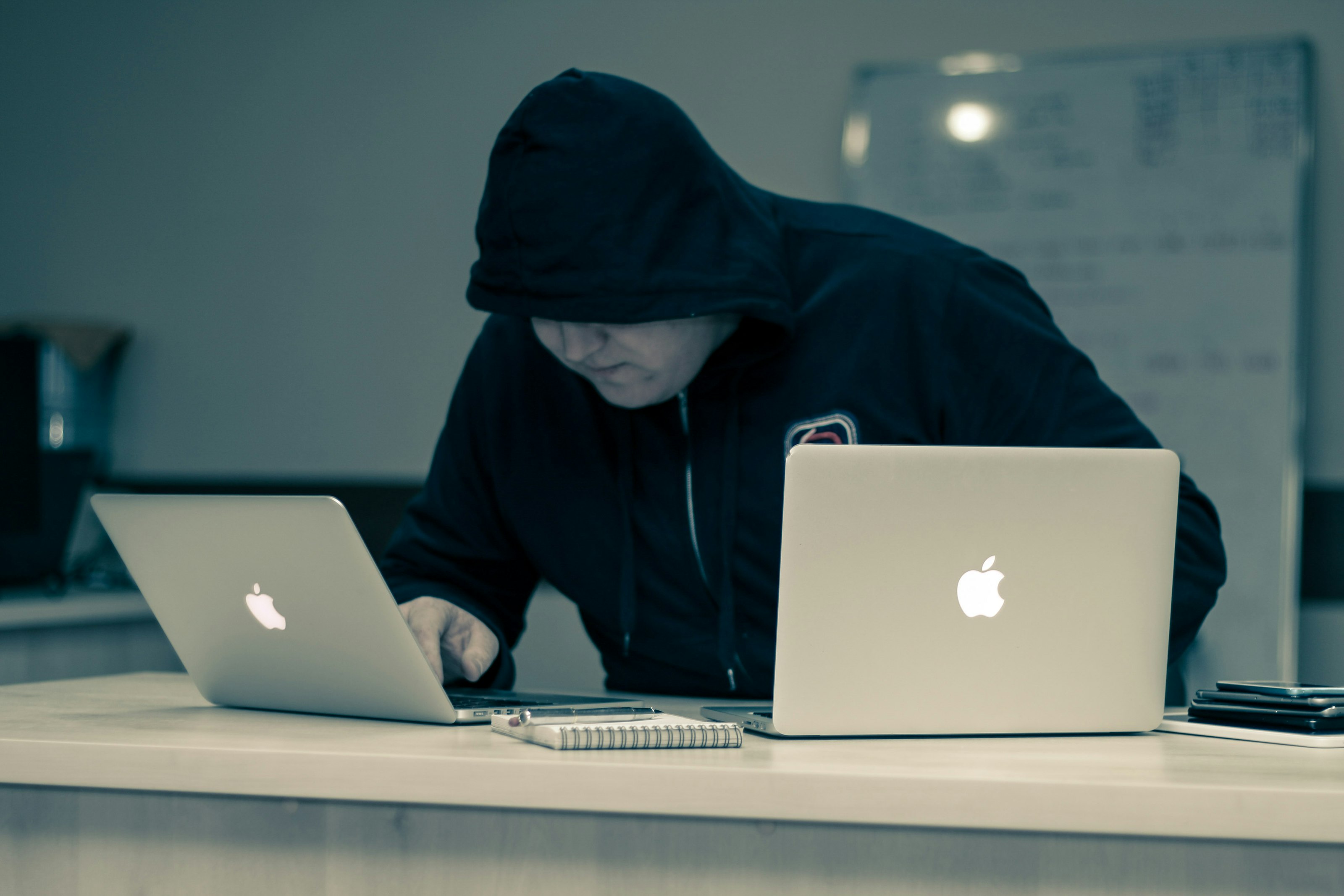 Person in black hoodie working with two MacBook laptops on a desk with a notebook and phone.